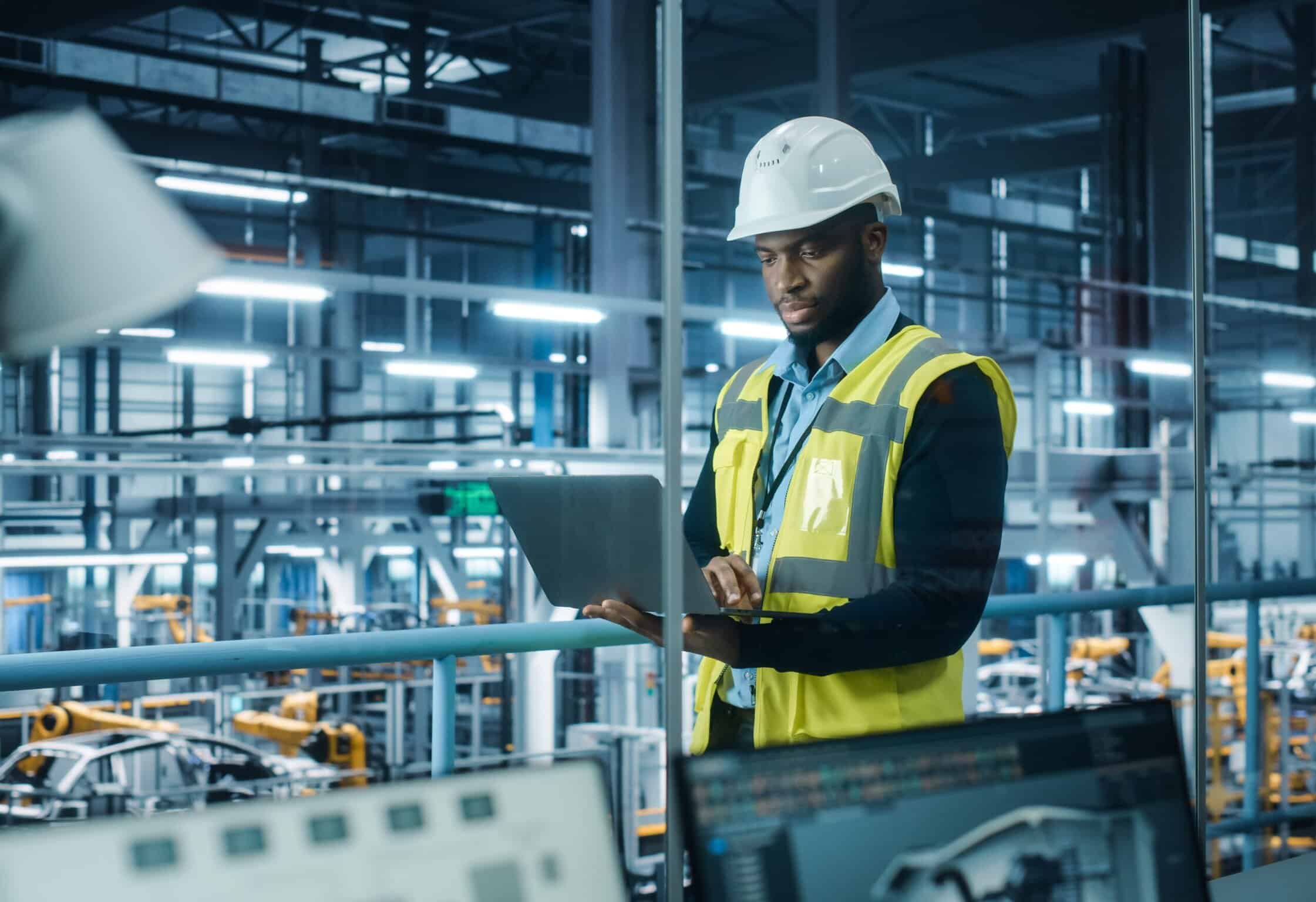 Industrial engineer inspecting machinery in manufacturing plant with laptop and safety gear.